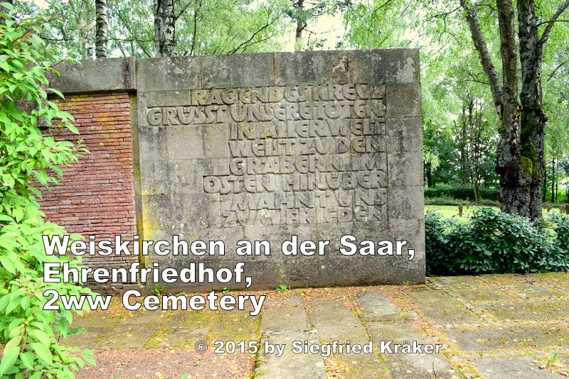 A view during a summer morning towards a stone wall with an engraved memorial plaque standing on the left side of the forecourt after the main entrance.